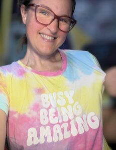 Sumara is a white woman wearing glasses. She is wearing a pink and yellow top which reads "Busy being amazing" and smiling happily.